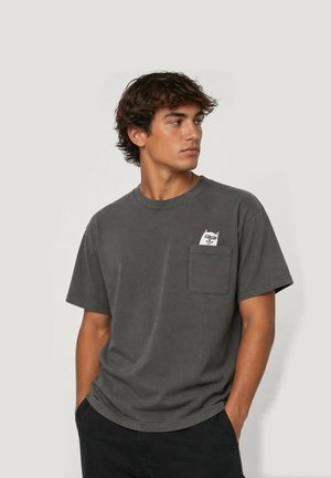 Joven con camiseta gris oscuro con un dibujo de gato asomándose desde el bolsillo del pecho, mirando hacia un lado, con las manos en los bolsillos de los pantalones negros.