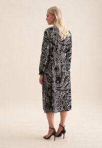 Schwarz-weiß gemustertes Kleid mit langen Ärmeln, tailliertem Schnitt und knielanger Länge. Kombiniert mit schwarzen High Heels, die eine glatte Stofftextur zeigen.