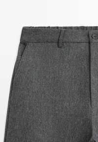 Pantalon gris en tissu texturé avec une poche latérale, fermeture à bouton et passants pour ceinture sur fond blanc.