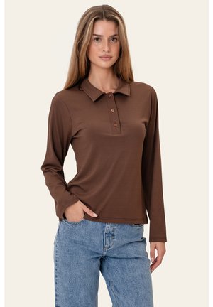 Kvinde med langt blondt hår iført en brun langærmet polo og lyseblå jeans, poserer med den ene hånd i lommen.