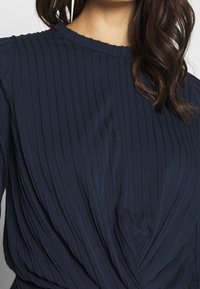 Mörkblå ribbad tygblus med texturerade veck, rund halsringning och snodd design detalj framtill.