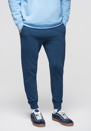 Mężczyzna ubrany w granatowe joggery, białe skarpetki i granatowe trampki z białymi sznurówkami, stojący z rękami w kieszeniach.