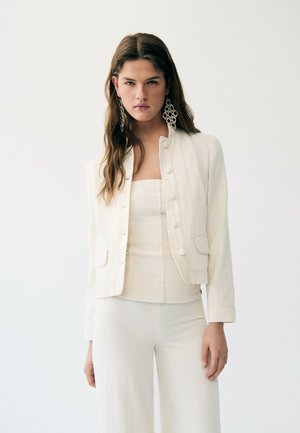 Femme aux cheveux longs bruns portant de grandes boucles d'oreilles géométriques, une veste blanche boutonnée, un top bustier et un pantalon large assorti.