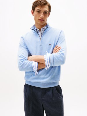 Hellblaues Sweatshirt mit einem Viertel-Zip, aus einem weichen Material gefertigt. Mit einem gerippten Saum und Bündchen, über einem weiß-blau gestreiften Shirt getragen.