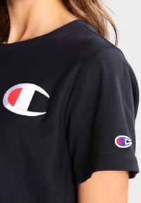 Camiseta de algodón negra con un gran logo circular bordado en rojo, blanco y azul en el pecho y un logo más pequeño en la manga.