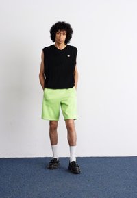Pull sans manches en tricot noir, shorts verts fluo, chaussettes blanches et chaussures noires. Design simple avec un col en V et un détail de logo.