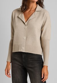 Donna che indossa un cardigan beige con bottoni, con polsini e orlo a coste, abbinato a jeans grigi scuri, su uno sfondo grigio uniforme.