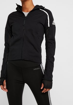 Femme portant une veste zippée noire Adidas avec des manches en mesh et des leggings noirs assortis avec des bandes blanches et le logo Adidas.