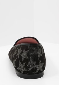 Zapato plano negro con un patrón de estrellas de purpurina plateada, tacón redondeado y forro interior rojo, visto desde atrás.