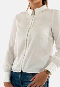 Blouse blanche à zip avec un col, des manches longues et des poignets élastiques. Fabriquée dans un tissu lisse et léger avec un éclat subtil.