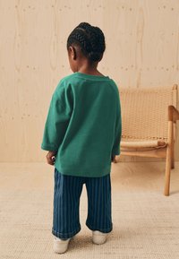 Grøn langærmet skjorte, løse blå stribede bukser, hvide sko. Barnet vender væk mod en let træbaggrund med vævet stol.