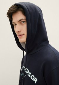 Felpa blu navy realizzata in cotone, con cappuccio regolabile e logo frontale. Texture morbida con una vestibilità rilassata e rivestimento interno grigio.