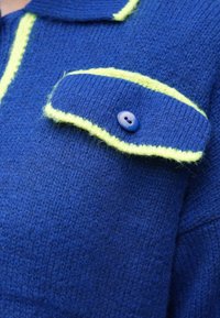 Niebieski dzianinowy sweter z kołnierzem, z jasnymi akcentami w kolorze limonkowej zieleni oraz zapinana na guziki kieszeń. Materiał jest miękki i puszysty.