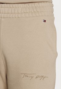 Beige tepláky s elastickým pasem, malá vlajka Tommy Hilfiger blízko kapsy a vyšitý text "Tommy Hilfiger" blízko stehna.