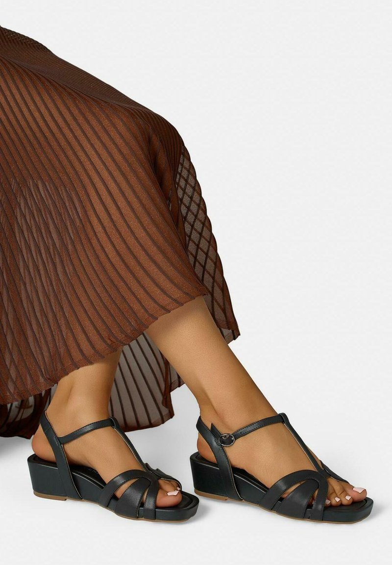 Pieds portant des sandales noires à talons bas avec lanières autour de la cheville, assorties à une jupe plissée marron sur un fond uni.