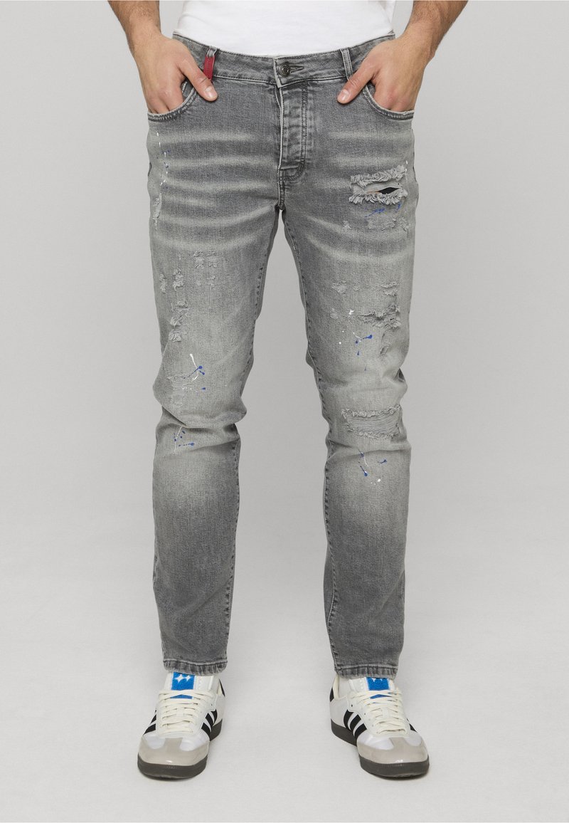 Man wearing faded gray distressed jeans with blue paint splatters and white sneakers with black stripes.