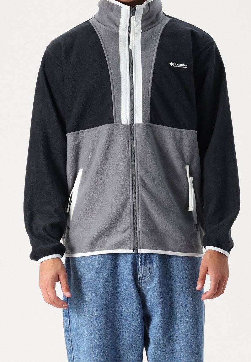 Grå och svart fleecejacka med hög krage, dragkedja fram och elastiska ärmslut. Har vita accentränder och sidofickor.