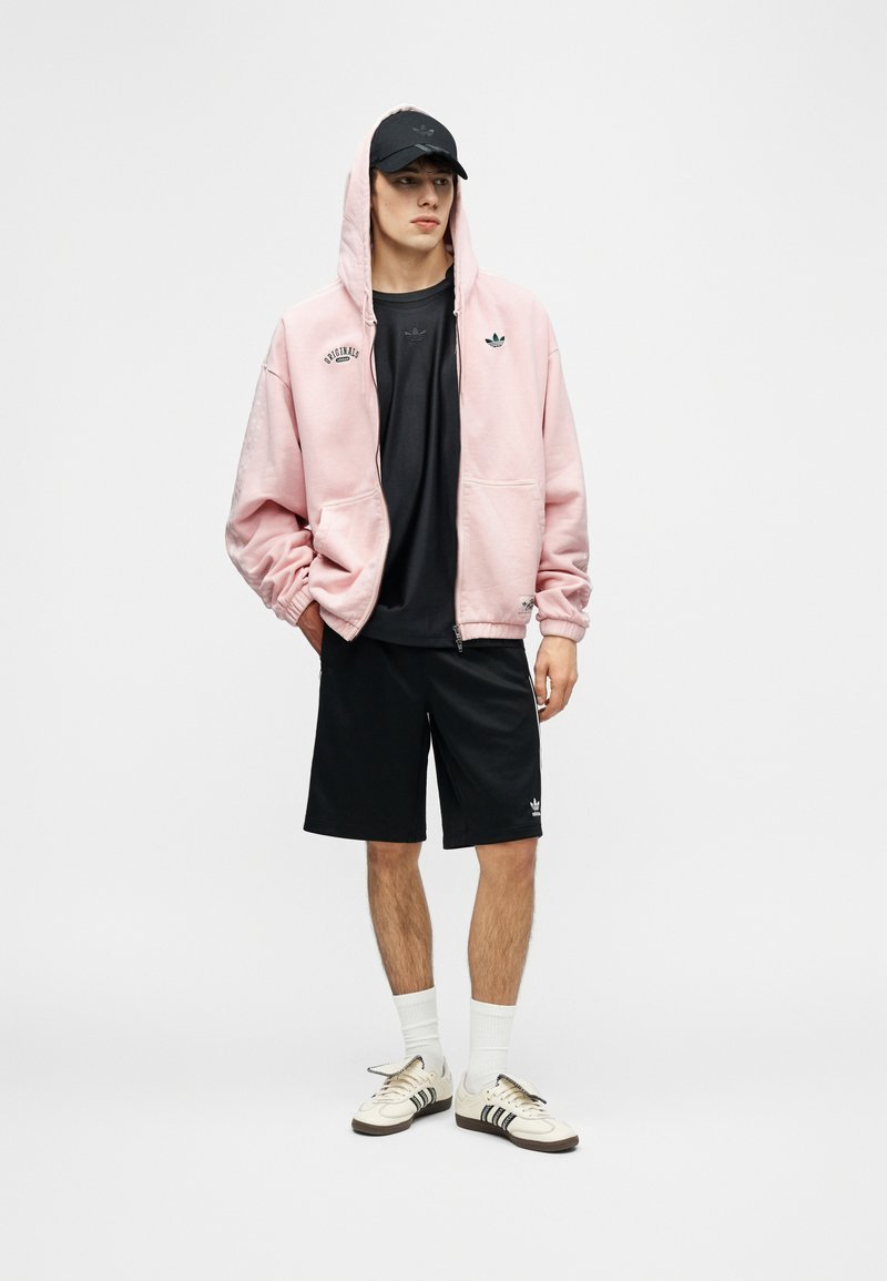 Jeune homme debout, portant un sweat à capuche rose zippé, une casquette noire, un t-shirt noir, un short noir, des chaussettes blanches et des baskets blanches avec des rayures noires.