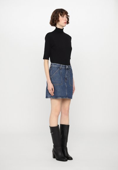 Mulher de pé de lado, vestindo uma blusa preta de gola alta, saia mini de denim com grandes bolsos frontais e botins pretos de salto alto até o joelho.