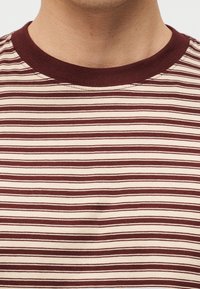 Κοντινό πλάνο ενός καφέ και μπεζ οριζόντια ριγωτού μπλουζάκι με μονόχρωμη καφέ στρογγυλή λαιμόκοψη, φορεμένο από ένα πρόσωπο.