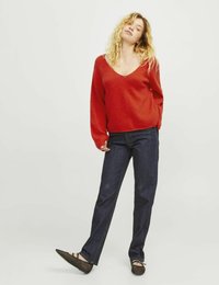 Roter, strukturiert gestrickter Pullover mit V-Ausschnitt und langen Ärmeln, kombiniert mit dunkelblauen geraden Jeans und schwarzen durchlöcherten Ballerinas.
