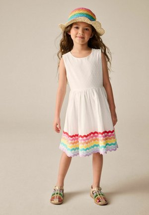 Junges Mädchen trägt ein weißes ärmelloses Kleid mit mehrfarbigem, wellenförmigem Saum, eine regenbogenfarbene Strickmütze und beigen Sandalen mit bunten Riemen.