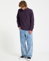 Mand iført en lilla sweater, acid-wash brede jeans og sorte sneakers, står med hænderne i lommerne mod en hvid baggrund.