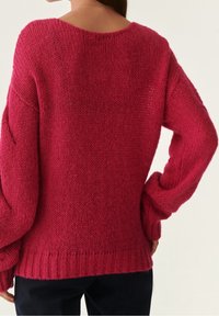 Suéter de punto rosa con un ajuste holgado, hombros caídos y dobladillo acanalado. La textura es suave y gruesa, con puntos abiertos.