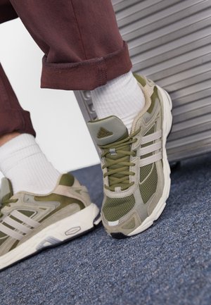 Baskets de sport vertes et grises en matériaux mesh et daim, avec une semelle légère et un logo à trois bandes, associées à des chaussettes blanches et un pantalon rouge.