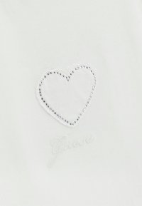 Camiseta blanca de algodón con un contorno de corazón texturizado hecho de tachuelas plateadas y texto bordado en la parte inferior.
