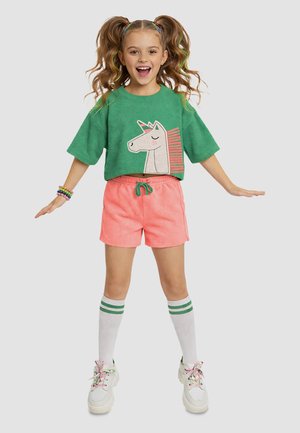 Ung pige med krøllede hestehaler iført en grøn unicorn sweatshirt, koralshorts, hvide knæstrømper med grønne striber og hvide sneakers.