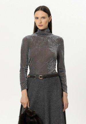 Woman with straight dark hair wears a grey velvet turtleneck, dark grey skirt with belt, and holds a small dark fur handbag against a white background.