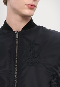 Črna bomber jakna z zadrgo, rebrastim ovratnikom in vezenim logotipom v temni niti. Gladko teksturo tkanine in prilegajoč kroj.