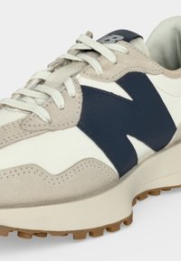 Gros plan d'une basket beige et blanche avec un logo latéral bleu marine, des lacets blancs et une semelle texturée marron clair.
