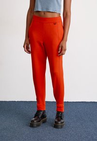 Persoon in felle oranje strakke broek en zwarte veterschoenen met platform die op een blauwe vloerbedekking staat tegen een witte muur.