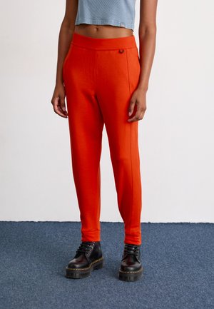 Person som har på seg knallorange tettsittende bukser og svarte snørestøvler med platå, står på blå teppe mot hvit vegg.