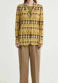 Blazer a fantasia con onde gialle, verdi e marroni, chiusura con un bottone, rever a punta, abbinato a pantaloni beige larghi.
