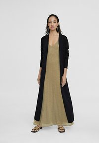 Sort langærmet cardigan over en metalisk guld maxi kjole; kjolen har en V-udskæring, mens sandalerne har et dekorativt mønster.