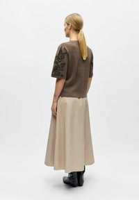 Femme avec queue de cheval blonde portant un haut marron à manches courtes avec broderies florales, une longue jupe beige et des bottes noires, debout de dos.