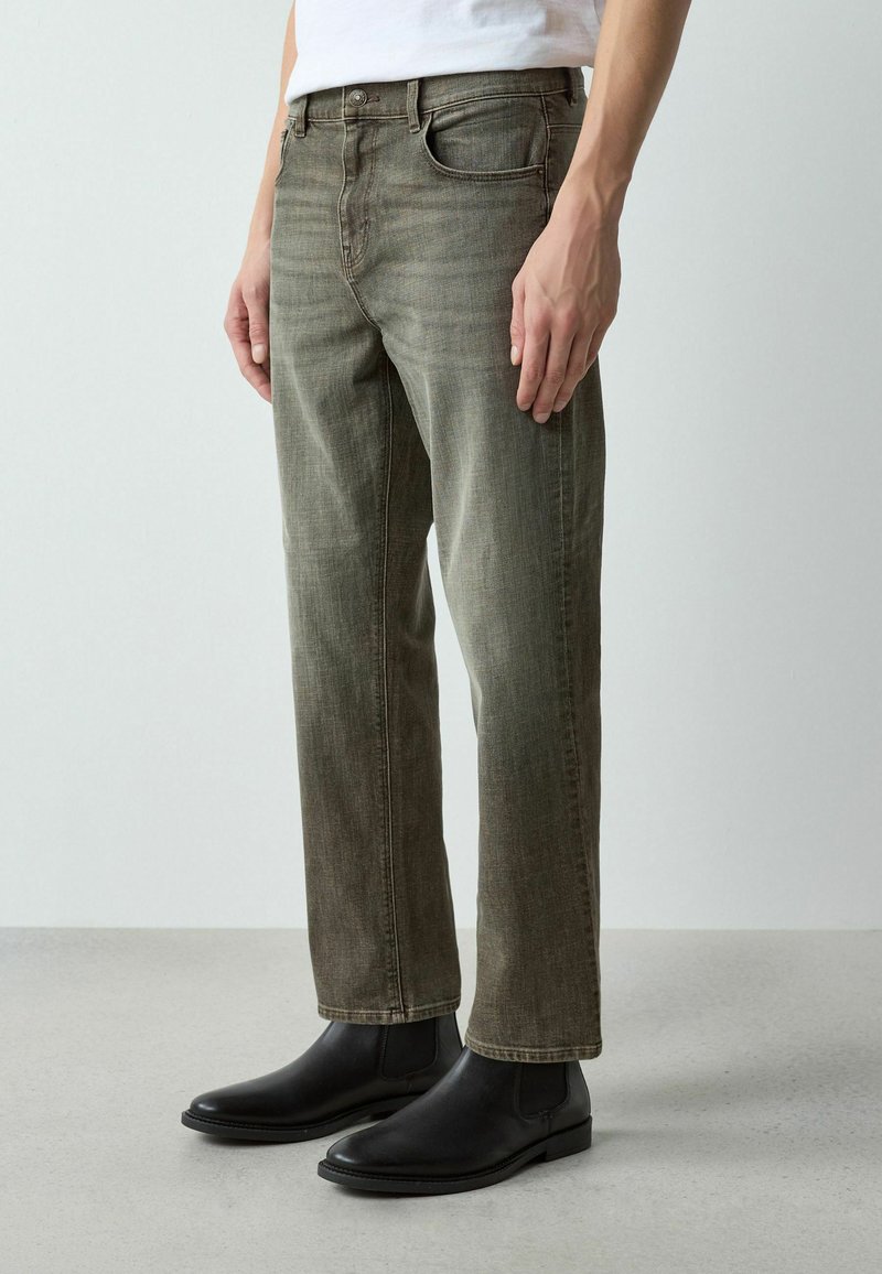 Homme portant un jean droit vert olive délavé, des bottines en cuir noir et une chemise blanche rentrée, debout sur un sol gris.