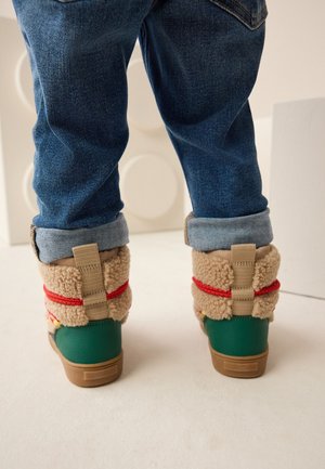 Enfant portant un jean bleu retroussé et des bottes d'hiver vertes avec une doublure polaire beige et des accents rouges, debout sur un sol clair.