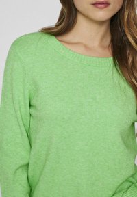 Svetlo zelena pletena jopica širokega kroga okoli vratu z dolgimi rokavi, ki se ponaša z mehkobo in diskretno rebrasto površino na manšetah.