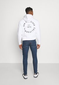 Sudadera blanca con estampado gráfico negro en la parte trasera, combinada con jeans azul oscuro. El modelo lleva zapatillas blancas y negras. Look simple y casual.
