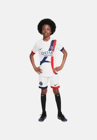 Witte jeugdvoetbaloutfit van Paris Saint-Germain met een blauw en rood ontwerp, voorzien van sponsorslogo's, zwarte shorts en knielange zwarte sokken.