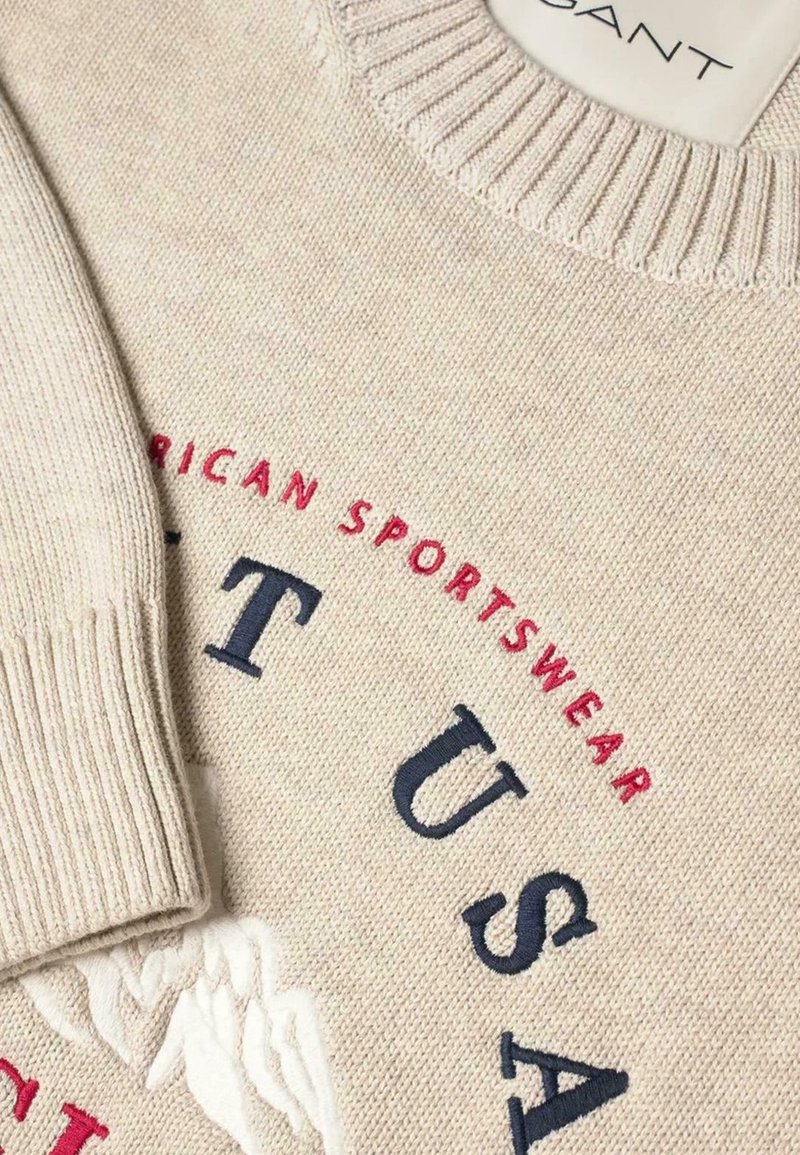 Maglione lavorato a maglia color crema con colletto e maniche a coste, testo ricamato "AMERICAN SPORTSWEAR" in rosso e "ATUSA" in blu navy.