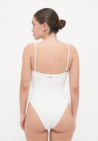 Maillot de bain one-piece blanc avec des bretelles fines réglables, un motif floral texturé, une coupe haute sur les jambes et un petit logo doré sur le dos.