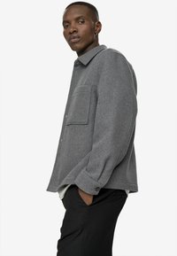 Cămașă-jachetă dintr-un amestec de lână gri, cu guler, un buzunar frontal, închidere cu buton și manșete răsucite, purtată peste o cămașă albă și pantaloni negri.