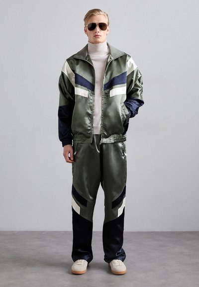 Veste de survêtement en satin vert olive avec des accents bleu marine et blanc, associée à un pantalon assorti. Comprend des poches zippées et un col haut.
