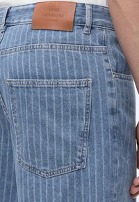 Οπίσθια όψη μπλε τζιν με λεπτές ρίγες, με μεγάλη πίσω τσέπη και δερμάτινη ετικέτα μάρκας "Mads Nørgaard Copenhagen" στη μέση.