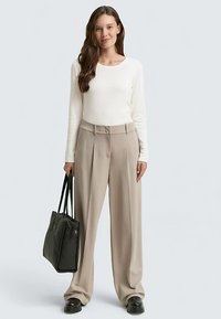 Kremfarvet langærmet top kombineret med højtaljede, vide beige bukser. Modellen holder en sort tote-taske og har sorte platåsko på.